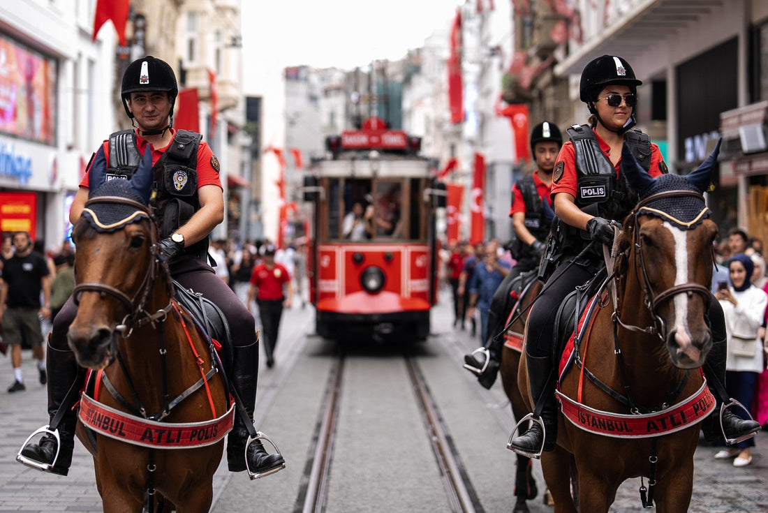 "Mounted police patrol with helmets and vests on brown horses, moving down Istiklal Avenue in Istanbul; a red nostalgic tram follows on tracks, showcasing security measures in place for tourists visiting the high-traffic Taksim square area in 2026."